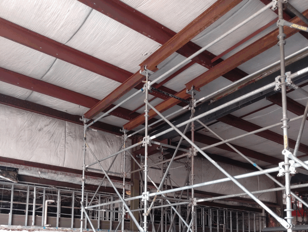Interior construction site showing scaffolding and rusted steel support beams beneath an insulated ceiling